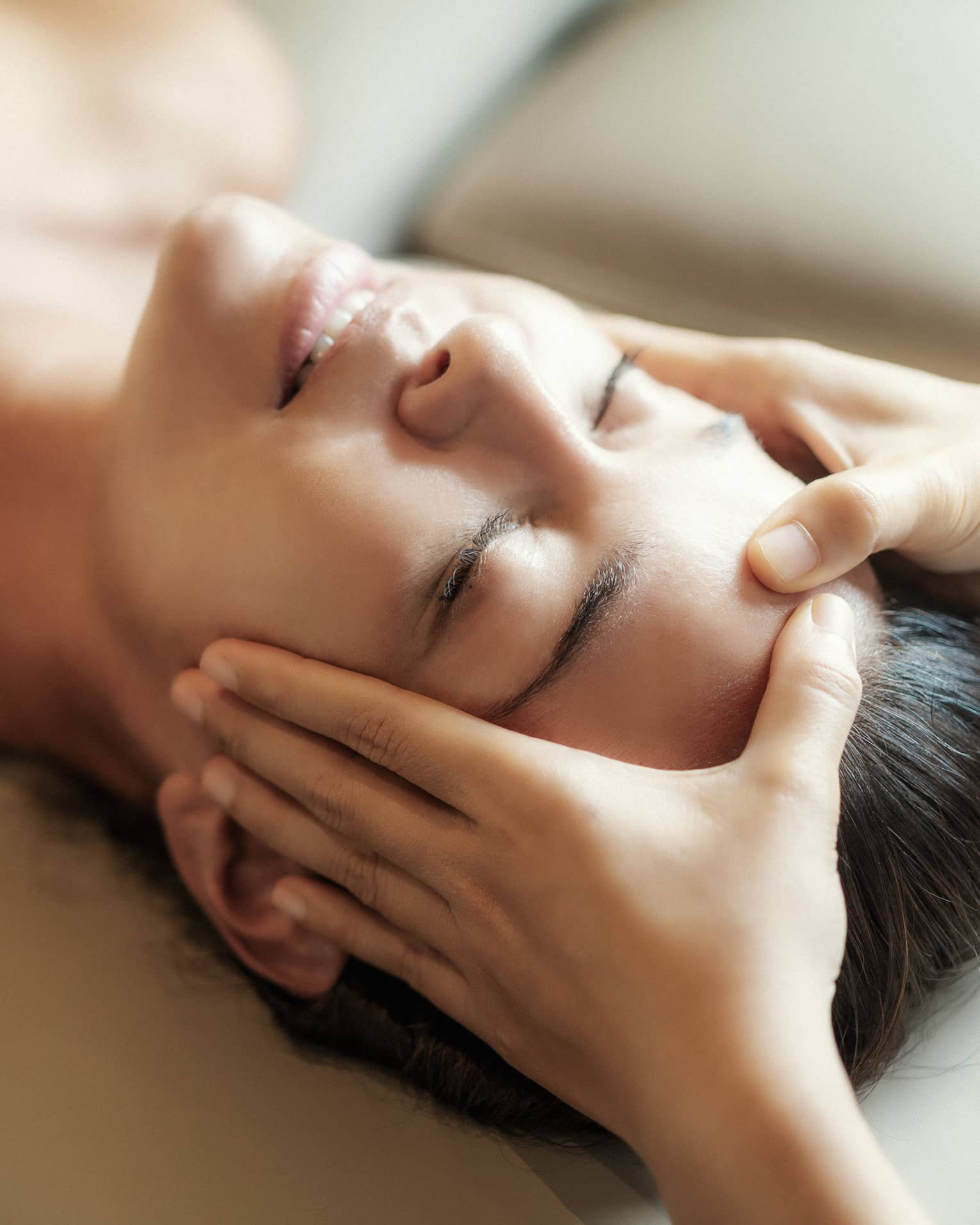 Spa facial, two hands rest on woman's forehead as she closes her eyes, lays under sheet on treatment table