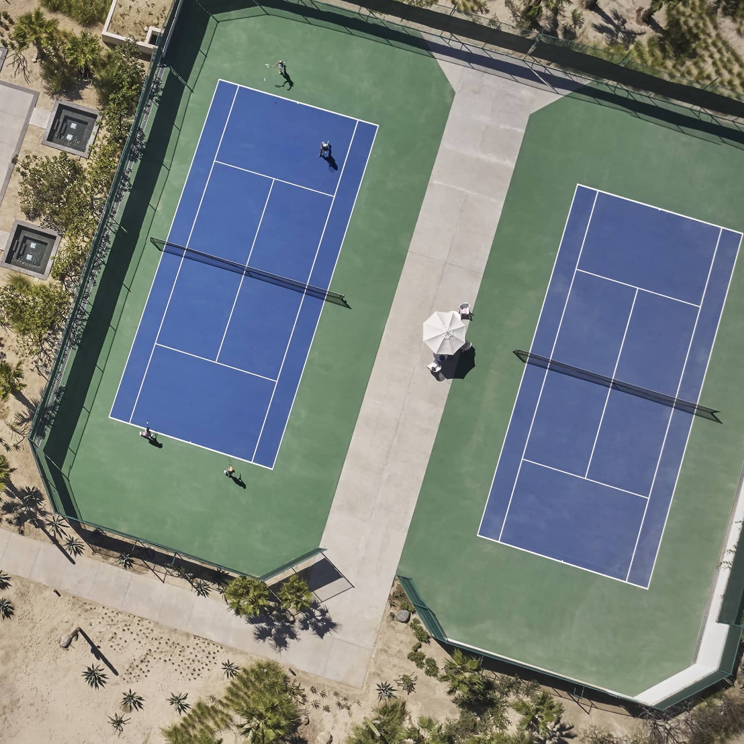 Aerial view of two green and blue tennis courts, one long rectangular pool and one blue basketball court surrounded by sand and greenery