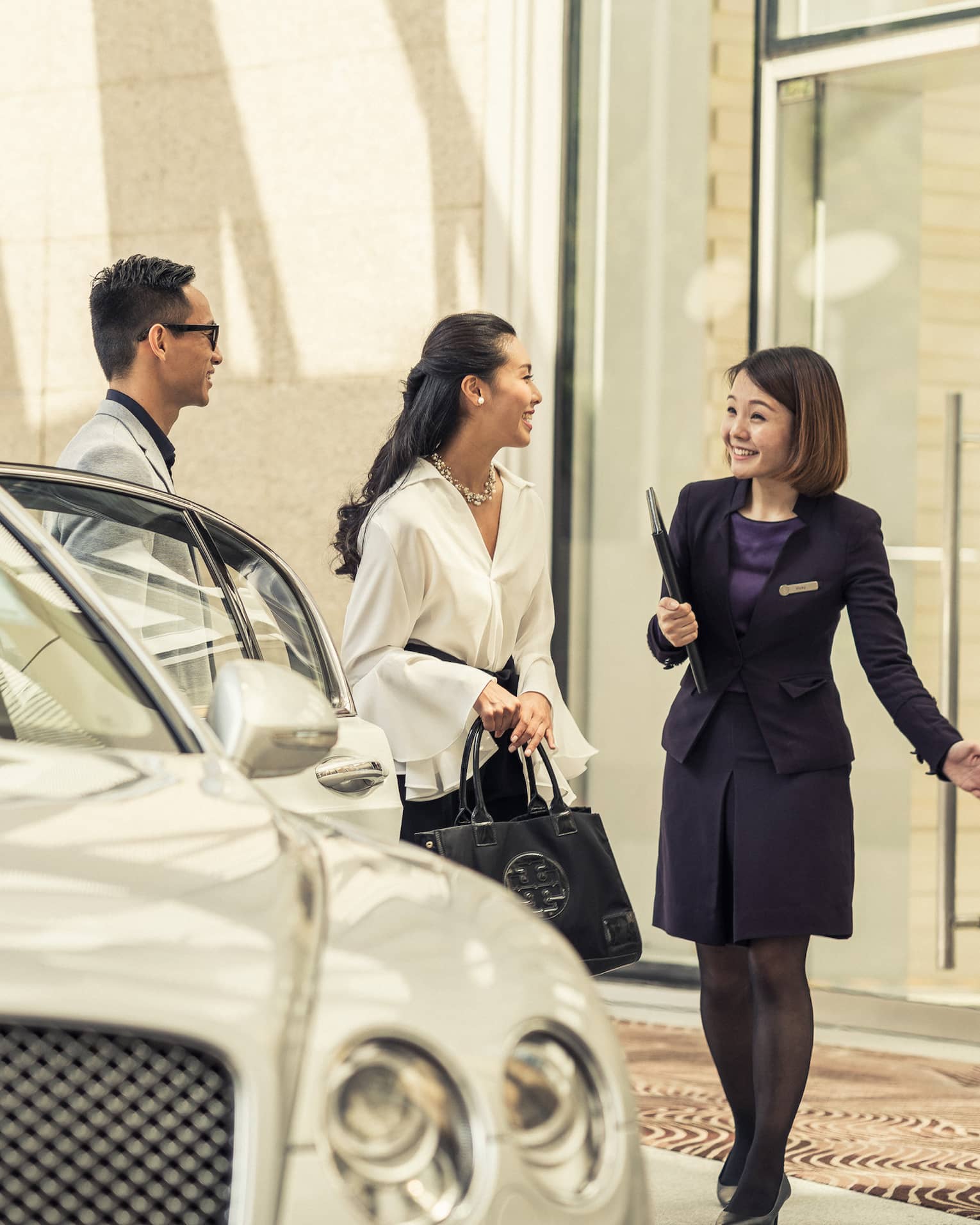 A person in a dark suit greets a couple near a luxury white car at the hotel entrance. The couple appears enthusiastic and engaged in conversation.