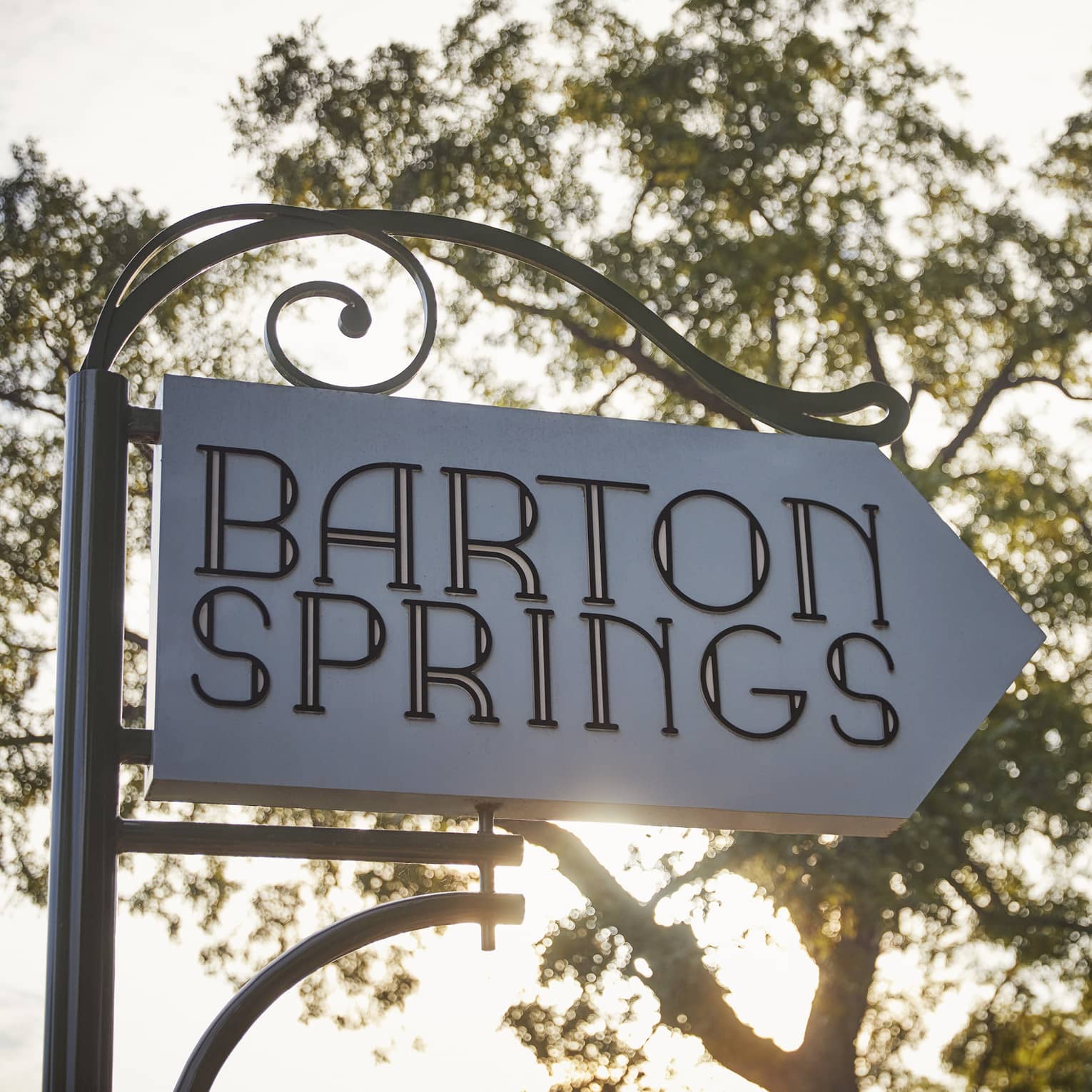 A white sign with a large tree and the sun behind it that reads "Barton Springs"