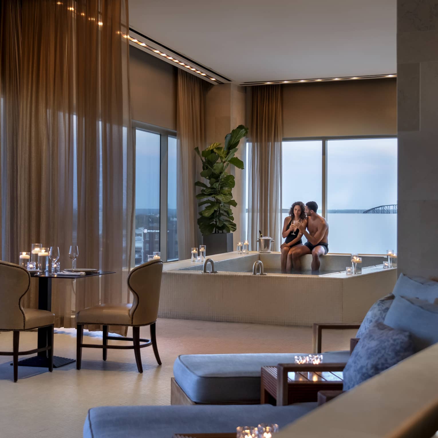 A couple enjoying a candlelit hot tub in a luxury hotel spa, with elegant seating, soft lighting and large windows offering a view of a bridge in the distance