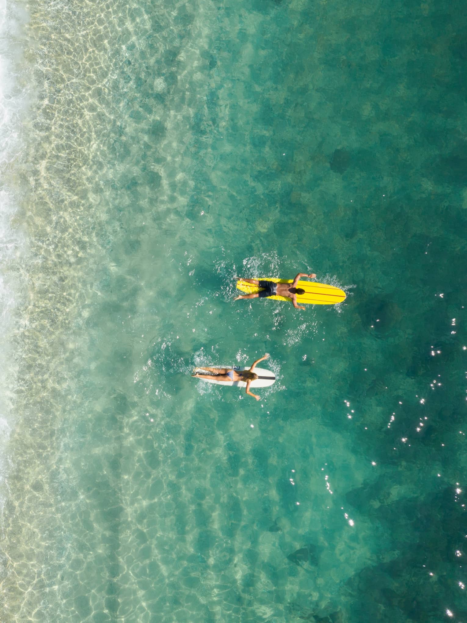 Bird’s-eye view of two people paddling their boards on their stomachs as frothy waves break on the beach behind them.