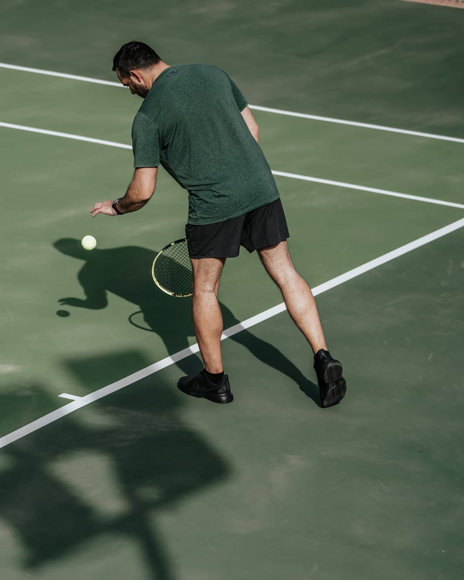 Man on tennis court bouncing tennis ball with left hand, preparing to serve
