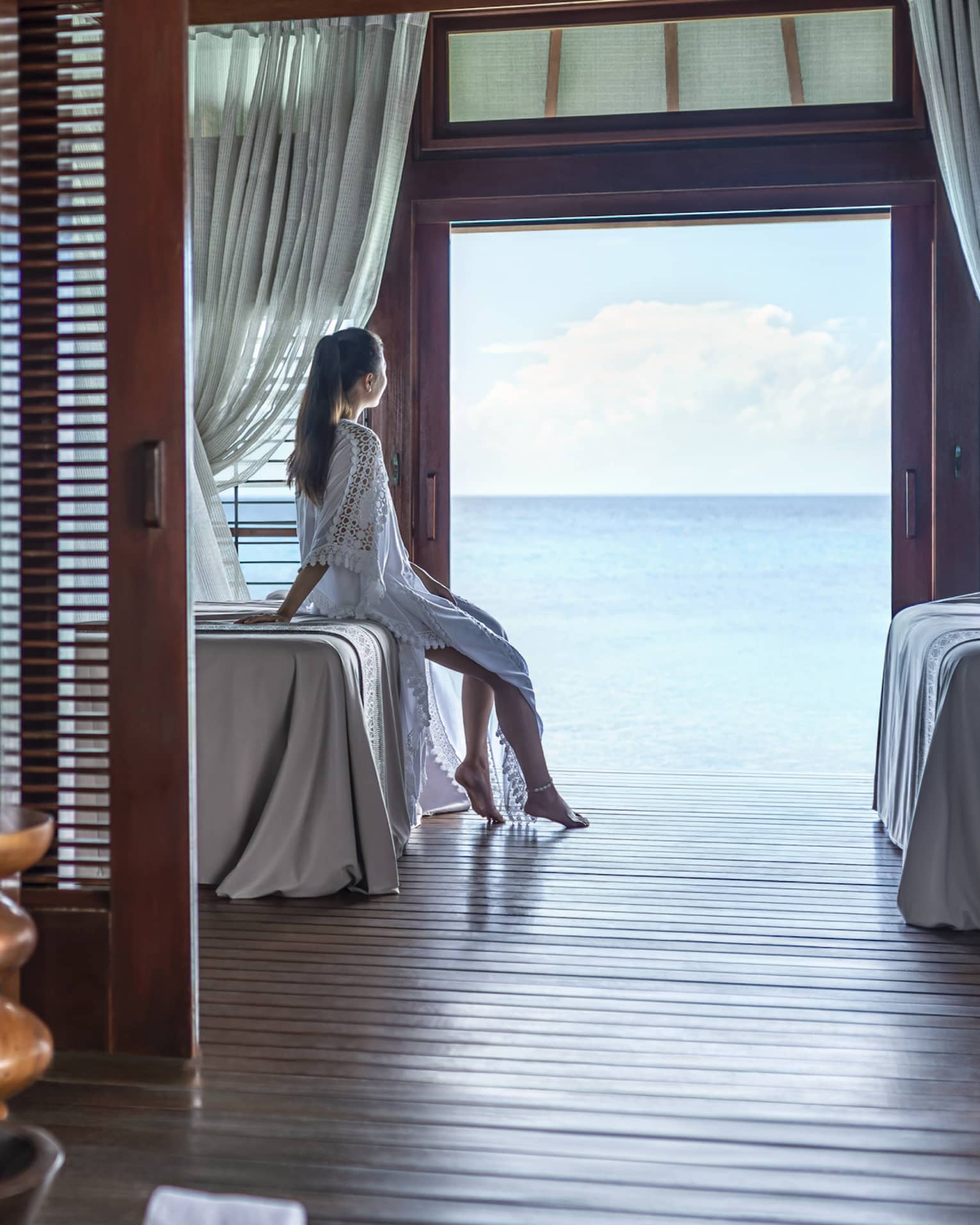 A woman leans on a massage bed on the far side of a spa treatment room and looks out through a large doorway onto the vast blue ocean