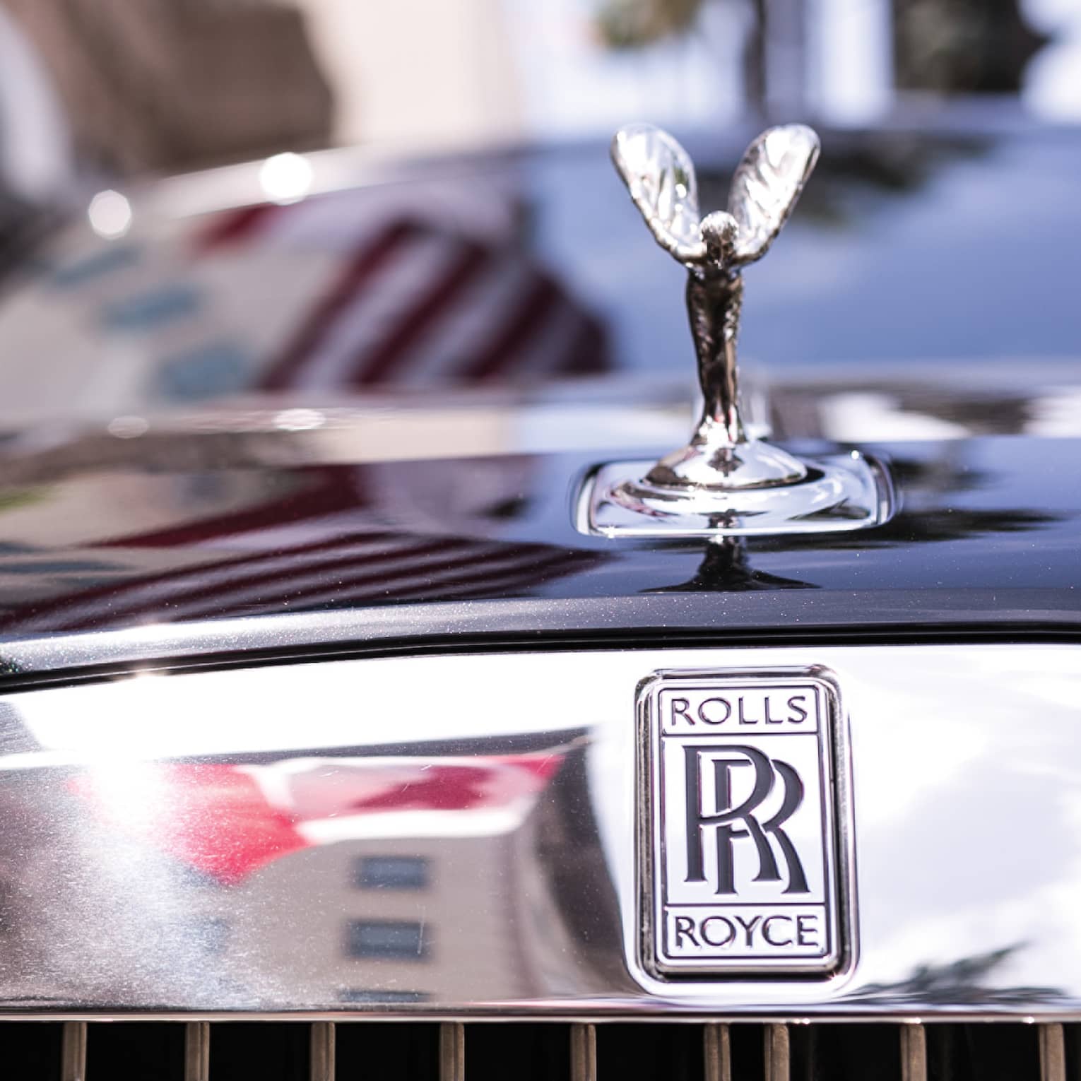 Close-up view of a winged silver ornament on the hood of black Rolls Royce, double-R logo on reflective chrome above grille.
