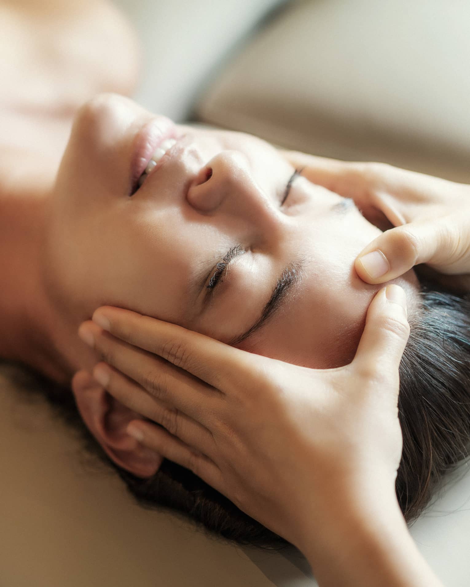 Spa facial, two hands rest on woman's forehead as she closes her eyes, lays under sheet on treatment table