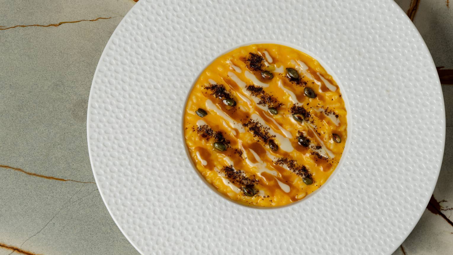 A shallow round bowl with a wide textured edge holds a orange-hued risotto preparation.