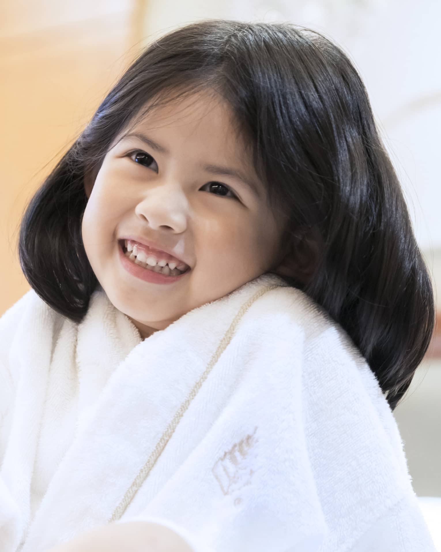 Smiling little girl wearing white bath robe and pink slippers sits on bed