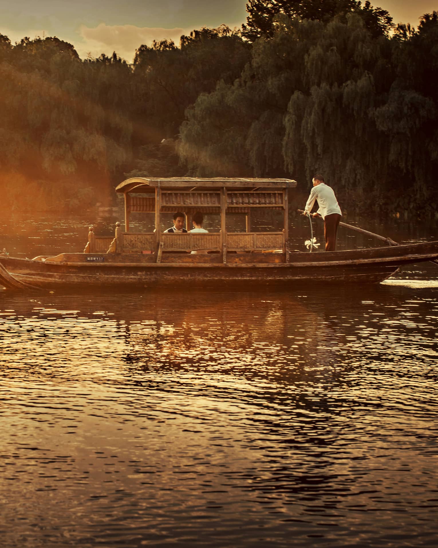 Man steers Chinese wooden rowboat on West Lake, into light of orange sunset