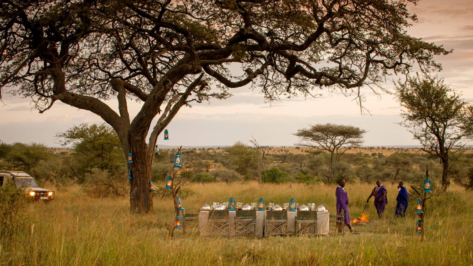 Three Maasai stand beside dining table and chairs, lanterns beside tree and jeep in Serengeti 