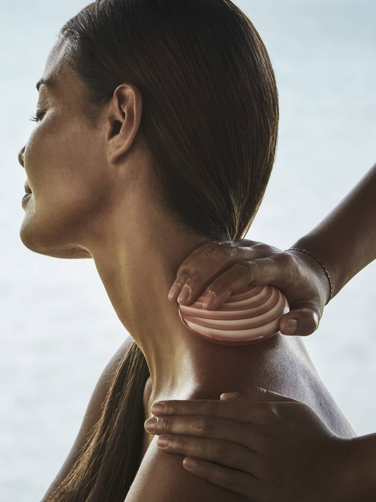 Side close-up of woman from shoulders up with head tilted to side as masseuse rubs seashell down her neck