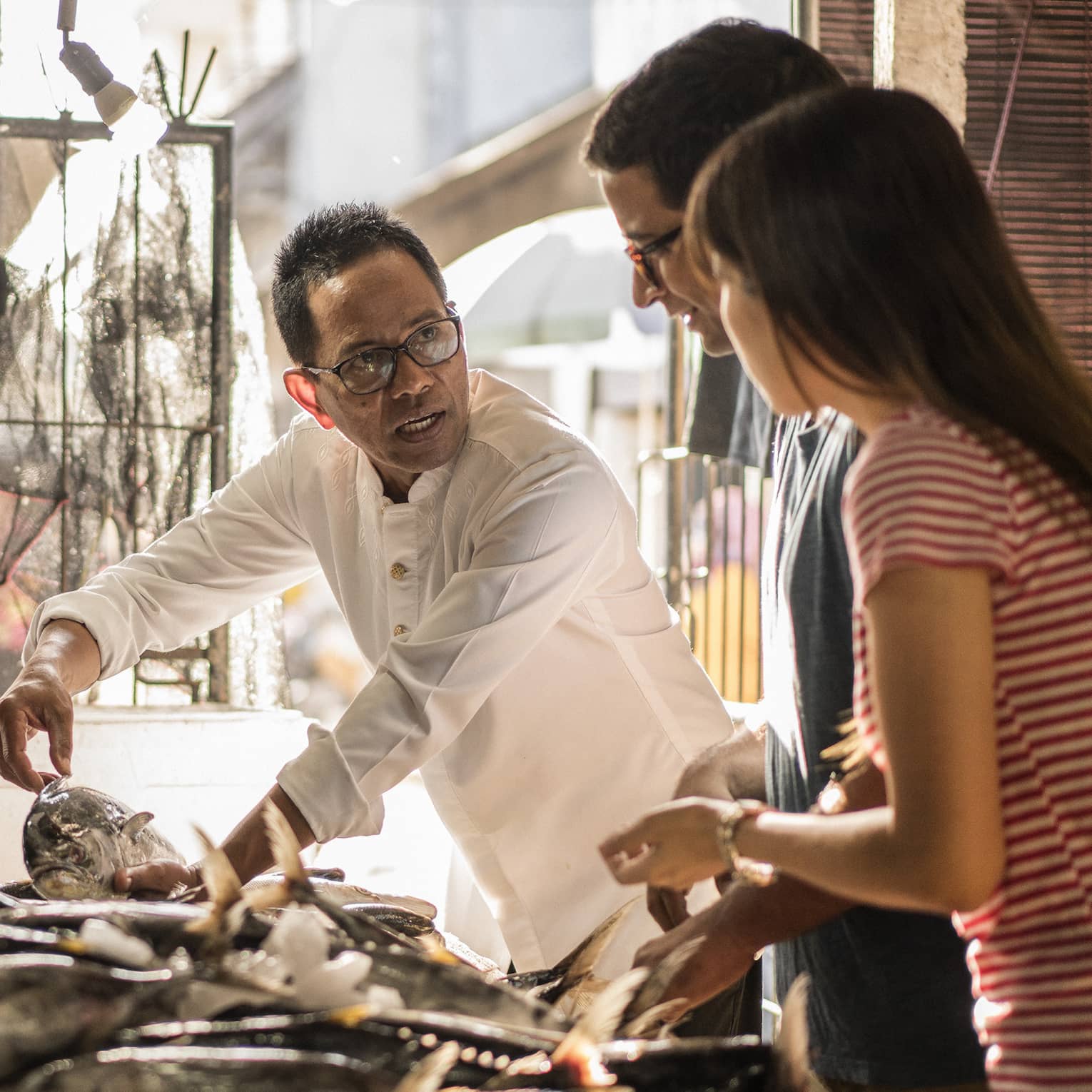A merchant gently holds a fish with two hands while talking to two guests as they all stand by an indoor market fish table.