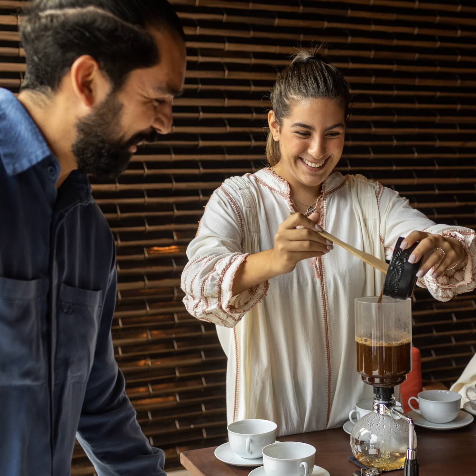 At a table set with white cups and saucers, two smiling guests watch as another pours coffee into a siphon coffee maker.