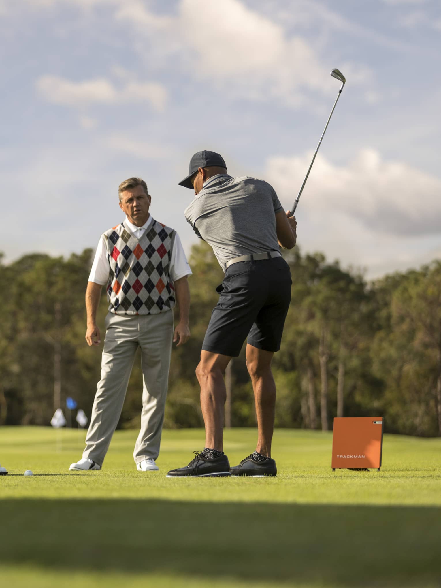 Against clear sky, one golfer looks on as another prepares to swing, a tray of balls and a stand of clubs nearby.