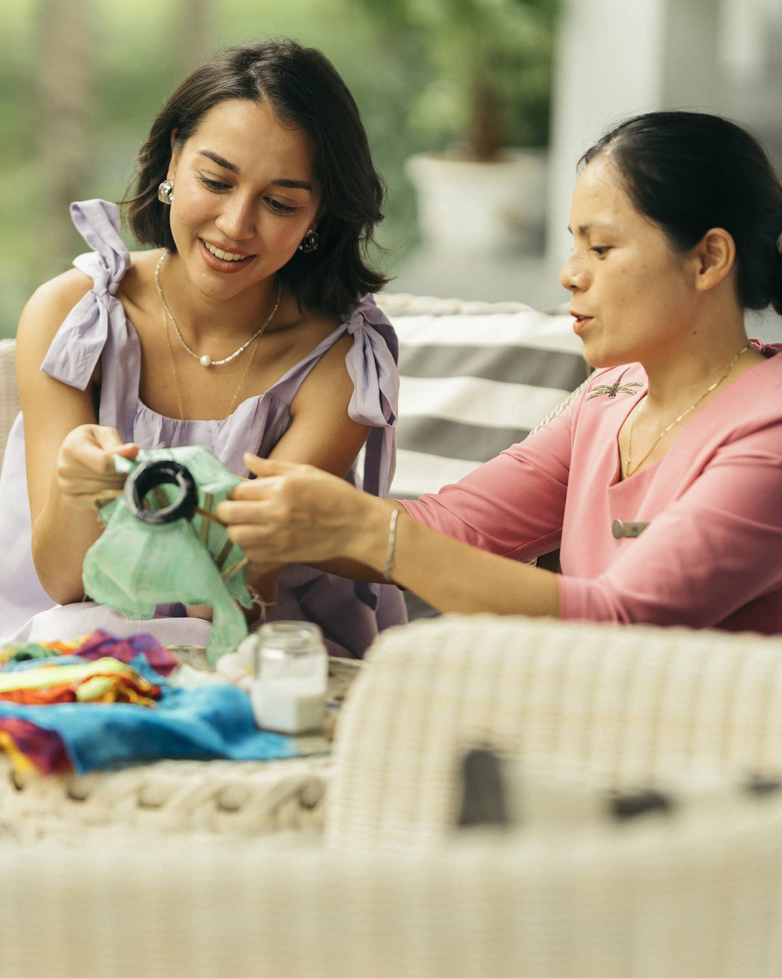 Two people engaged in a traditional craft activity, working together on fabric art while seated on a wicker patio set in a lush, green outdoor setting.