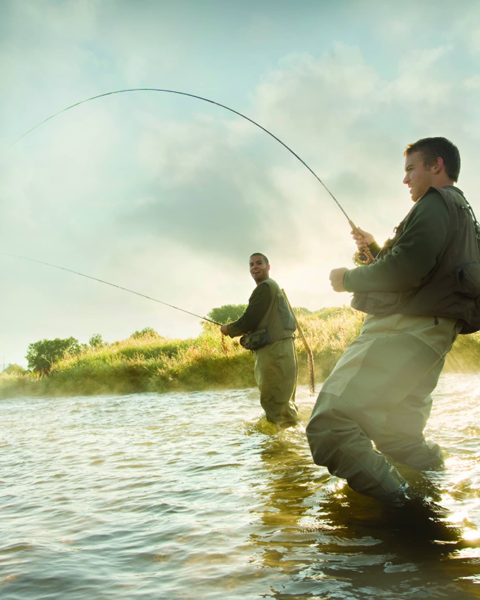 Two people fly fishing in a river, standing knee-deep in the water, surrounded by mist and greenery.