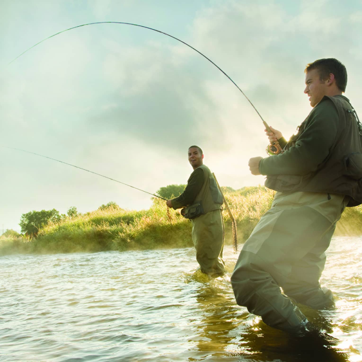 Two people fly fishing in a river, standing knee-deep in the water, surrounded by mist and greenery.
