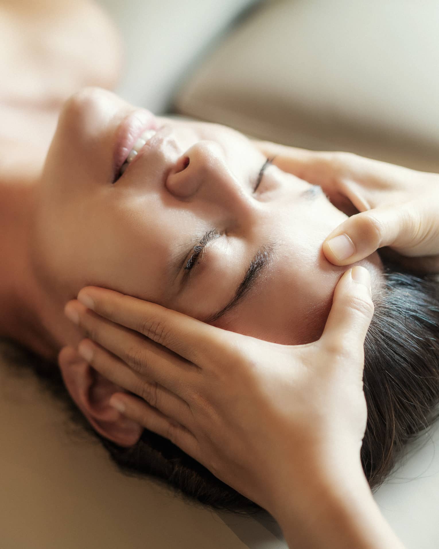 Spa facial, two hands rest on woman's forehead as she closes her eyes, lays under sheet on treatment table