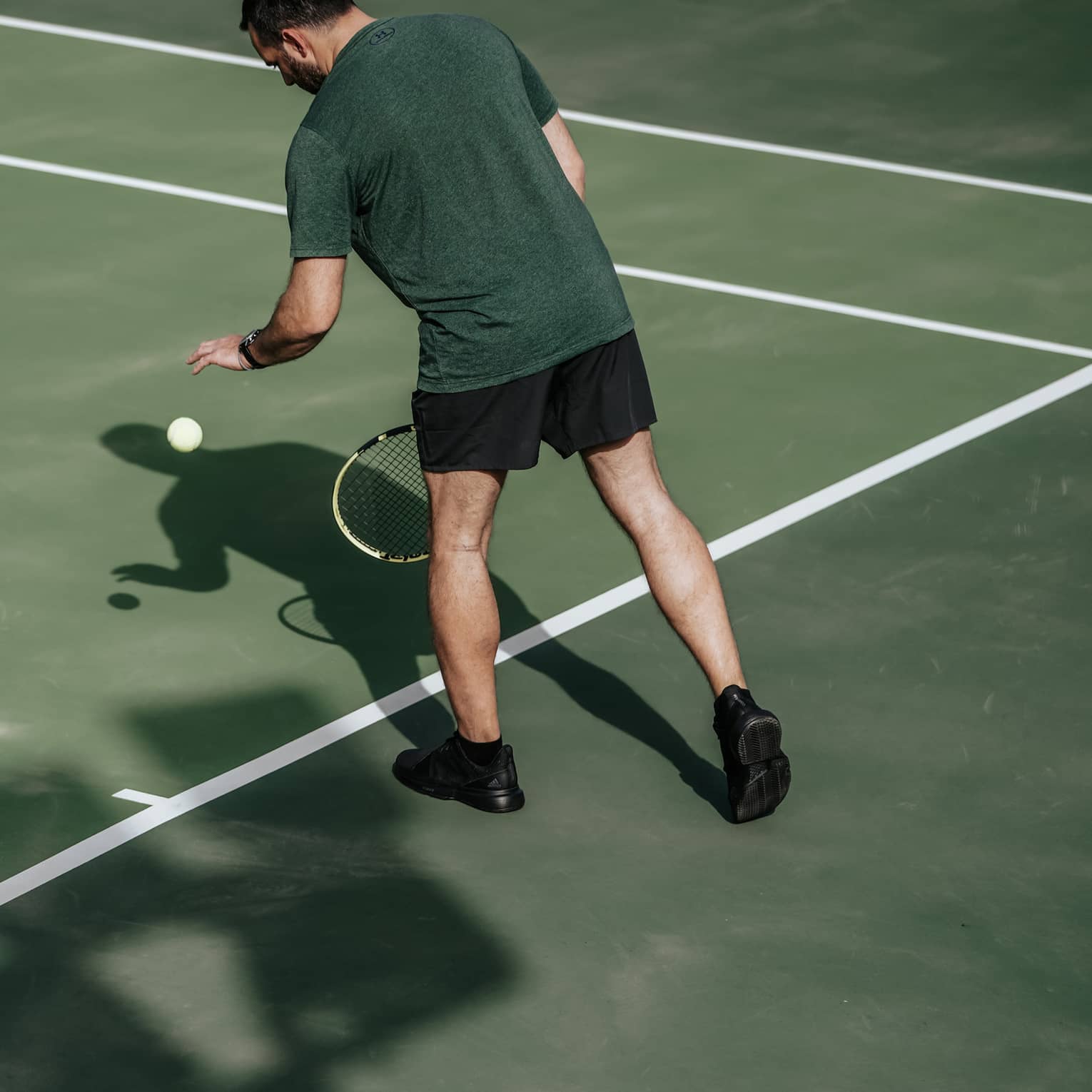 Man on tennis court bouncing tennis ball with left hand, preparing to serve