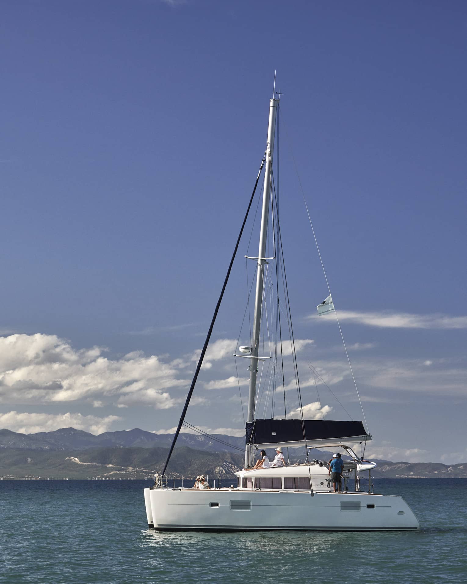 A catamaran on calm, blue water under a partly-cloudy sky with low green hills in the background and mountain ranges beyond.