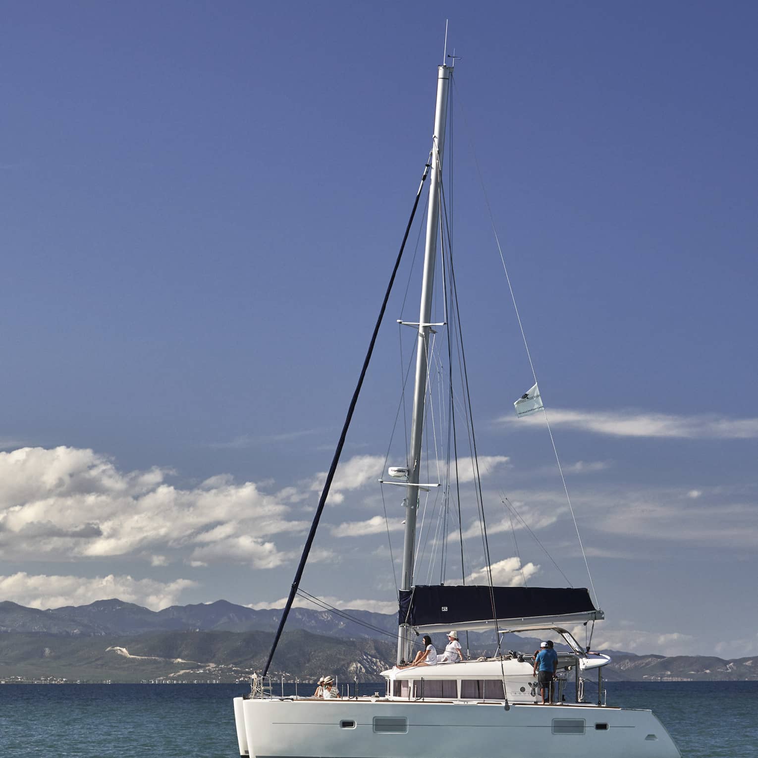 A catamaran on calm, blue water under a partly-cloudy sky with low green hills in the background and mountain ranges beyond.