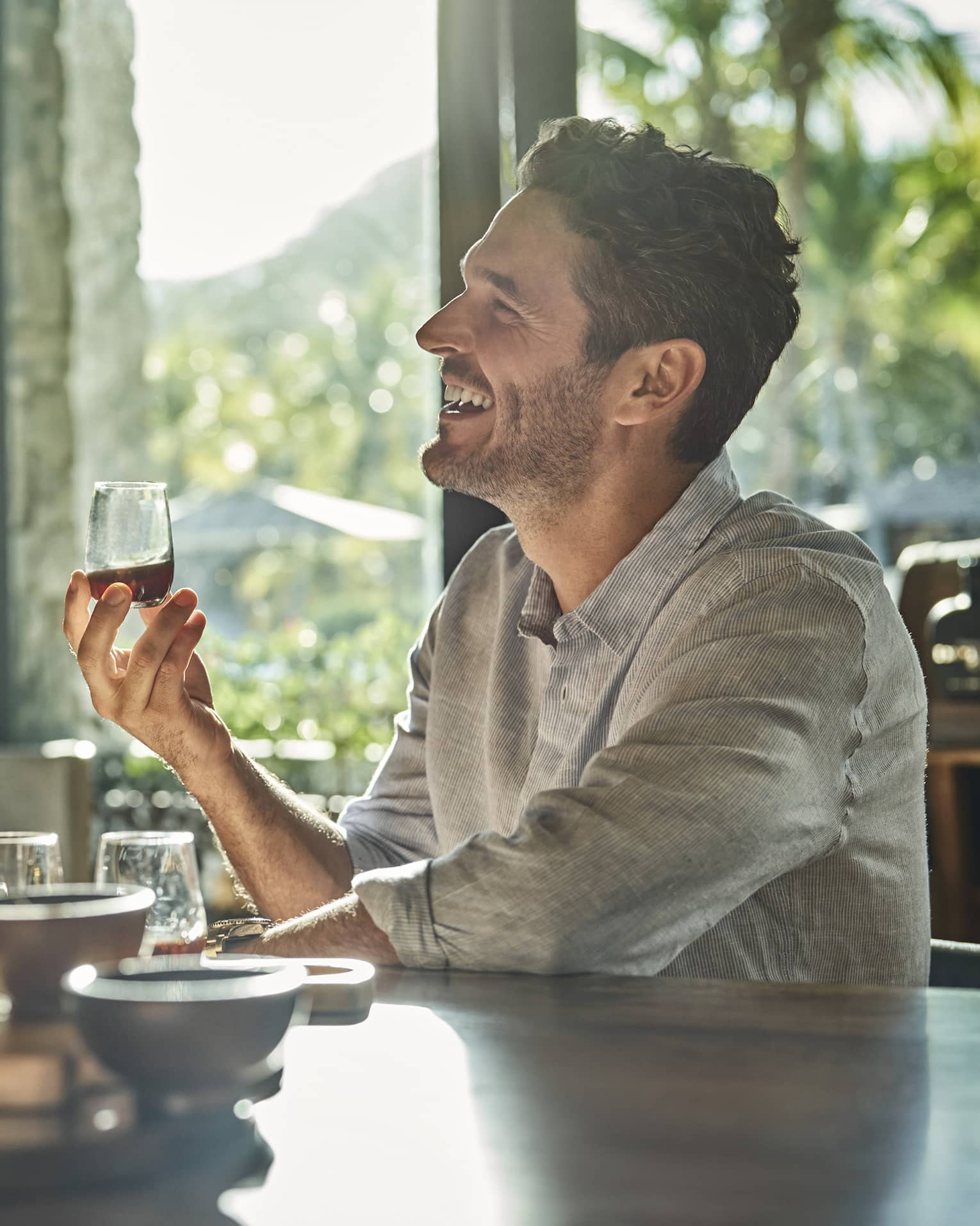 A man and a woman sit at a counter looking up and smiling and each holding a glass of rum with various other glasses and ingredients sit in front of them on the counter