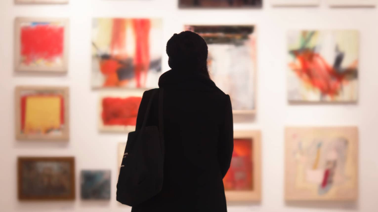 Silhouette of back of woman with hat bag looking at gallery wall with colourful art