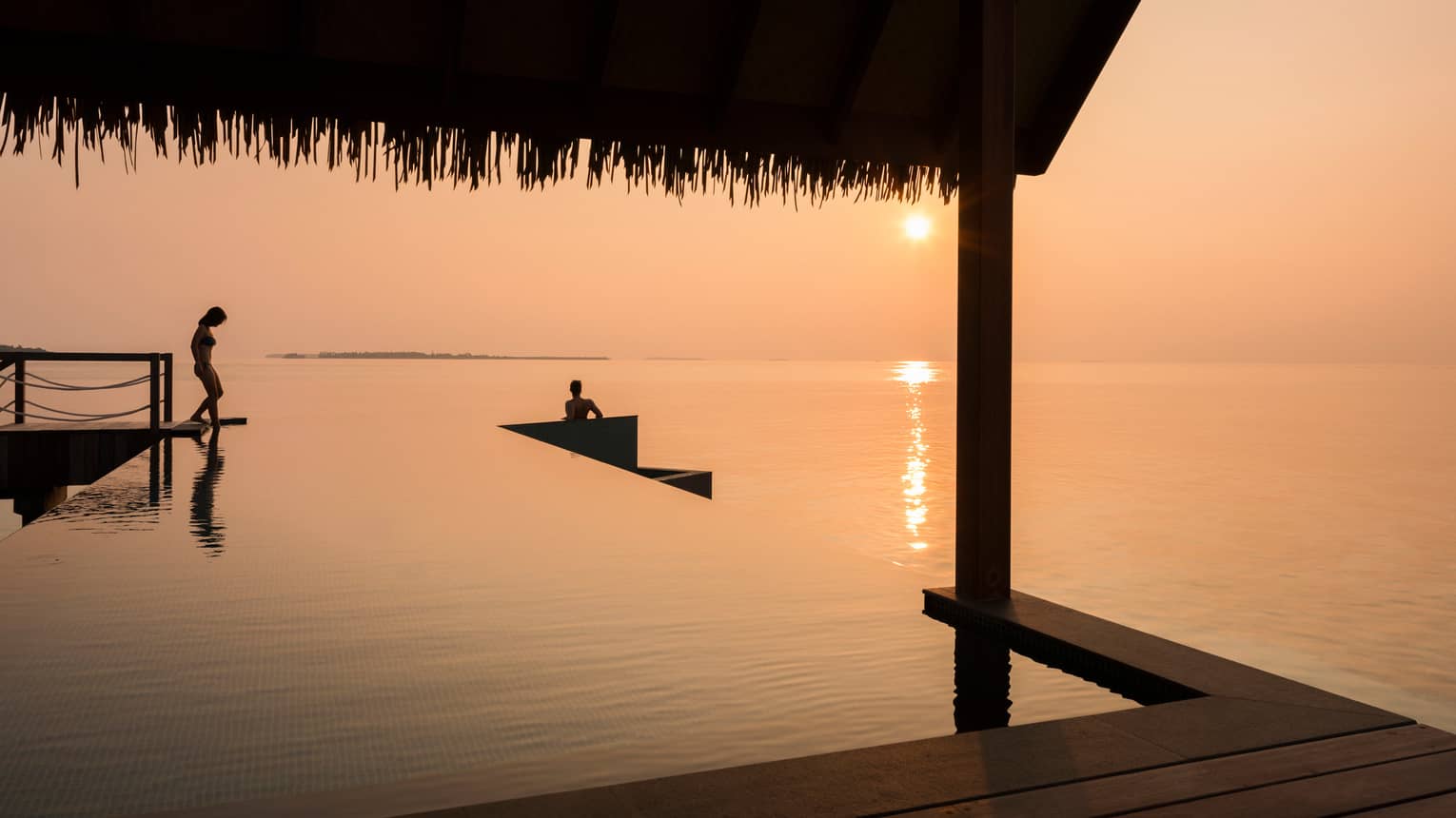 Orange sunset over ocean, silhouettes of people stepping into infinity pool under cabana