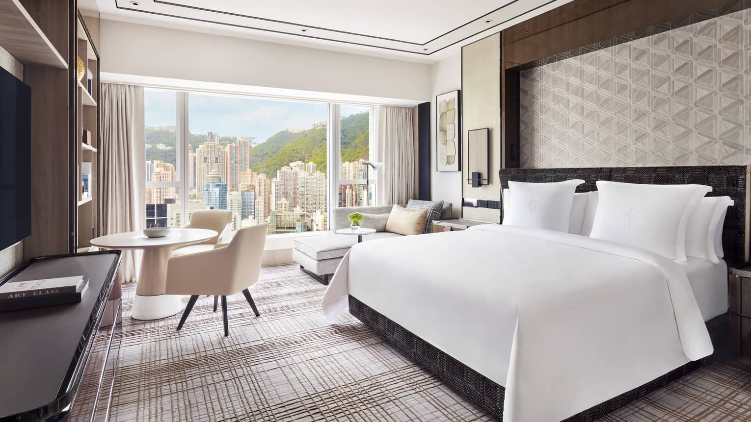 Modern hotel room with a large bed, geometric wall design, and a seating area next to a round table. Floor-to-ceiling windows offer a view of skyscrapers.
