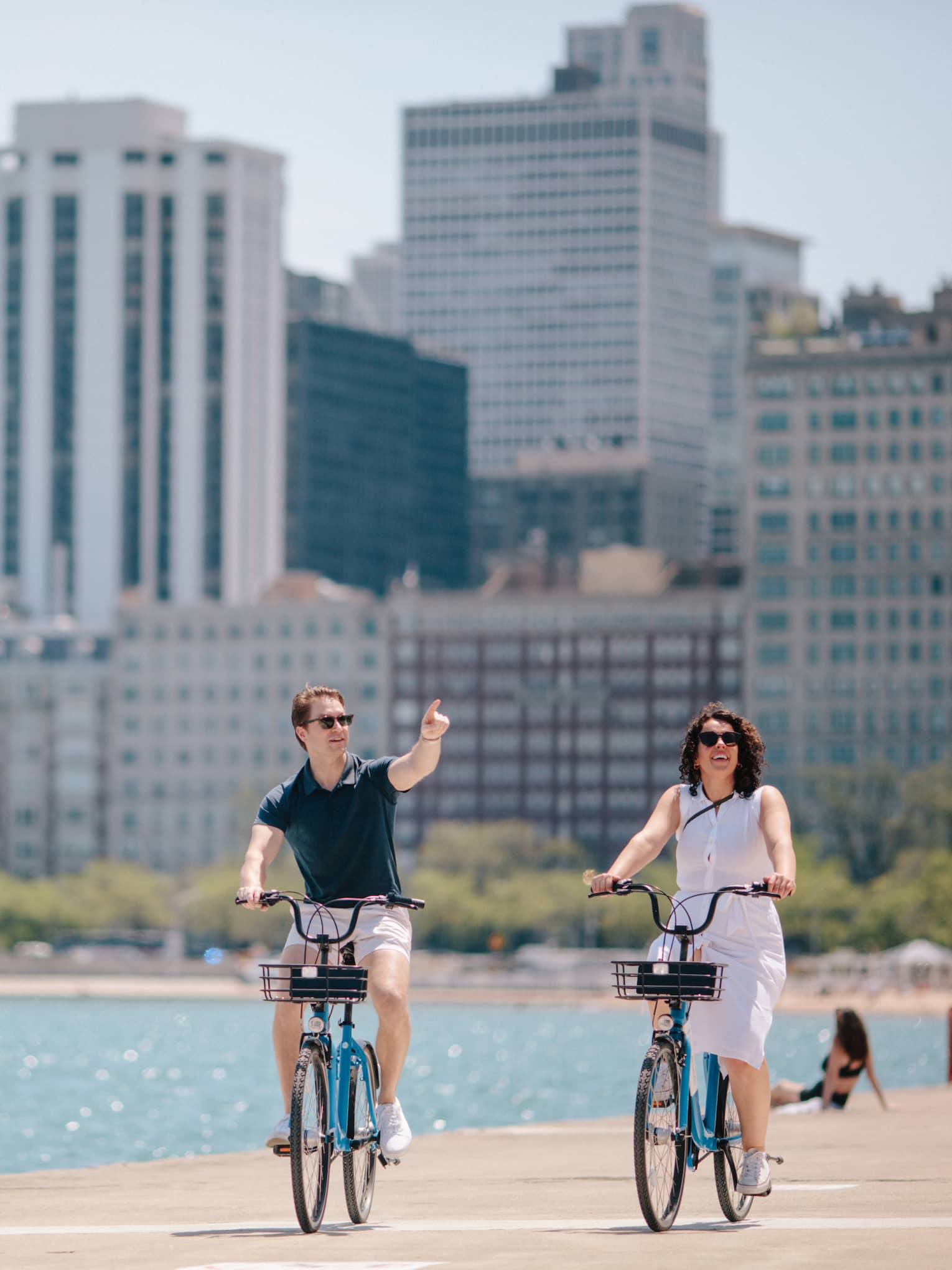 A man and woman cycling next to water with a city behind them.