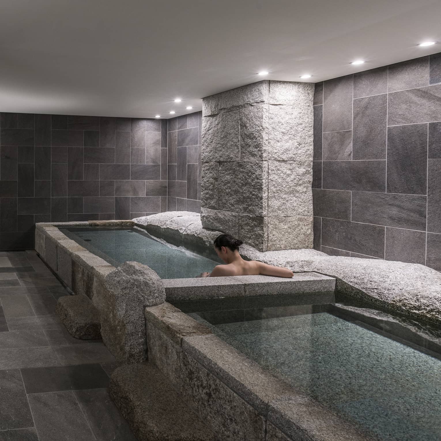 Back of woman's bare shoulders and she soaks in spa tub in tile, granite room