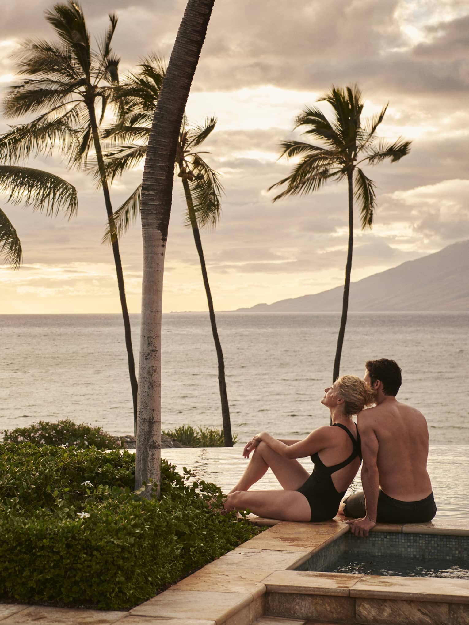 Man and woman lounge at pool's edge overlooking ocean in Maui