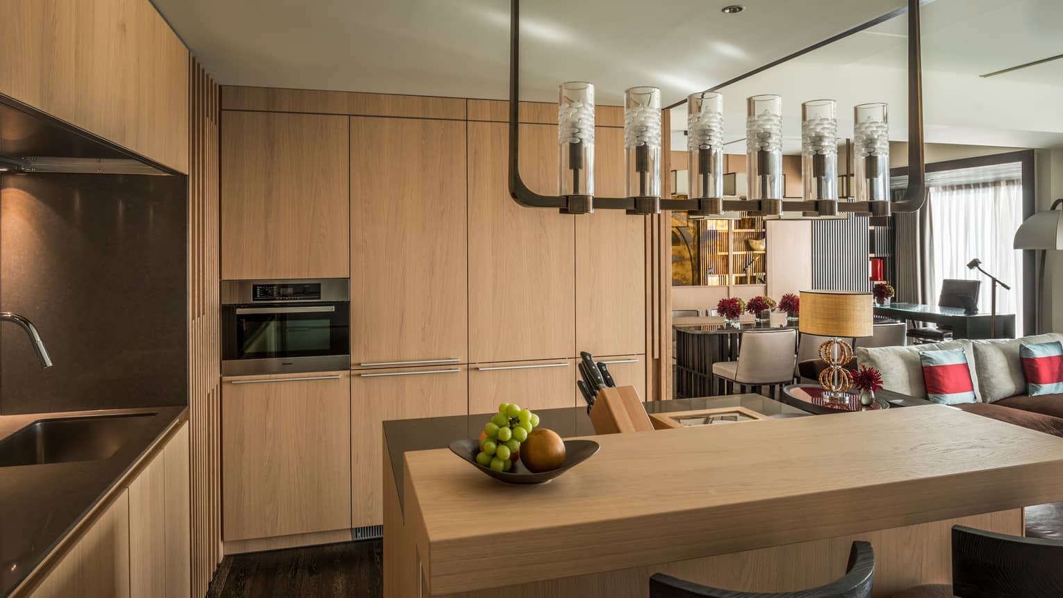 Modern kitchen area with light wood cabinetry, a built-in oven, sink and a central island with a fruit bowl, leading into a cozy Four Seasons living space