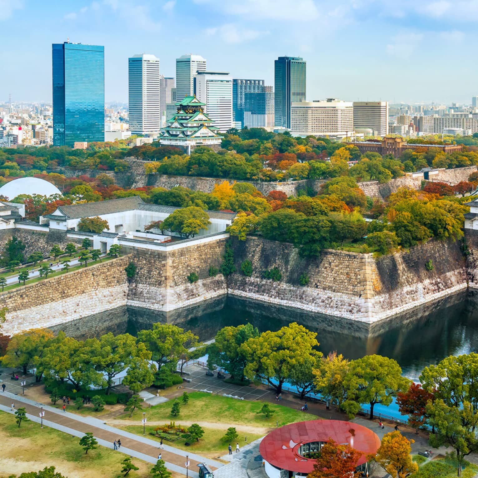 Osaka Castle rises amid lush autumn-coloured trees within a stone fortress surrounded by a moat and modern high-rises.