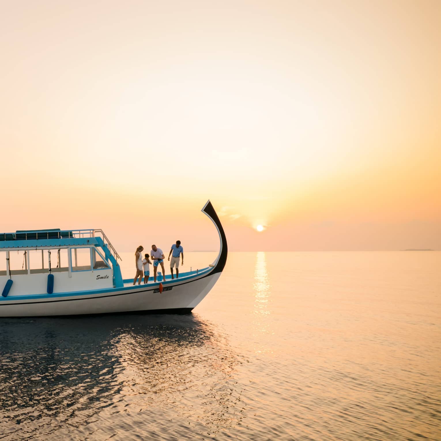 In sunset's golden glow, four people on the prow of a dhoni boat peer over the side as one pulls up a red fish on a line.