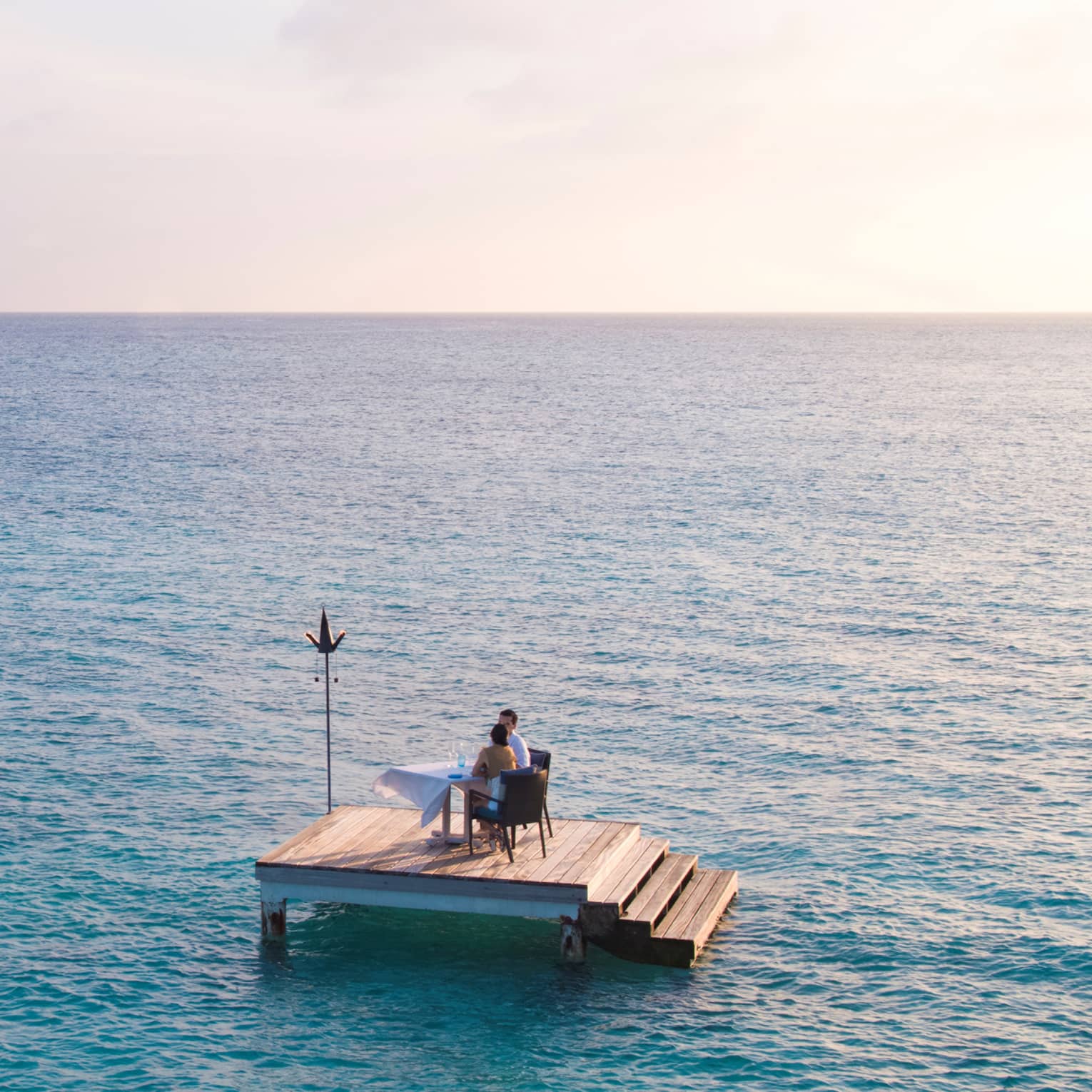 Man, woman dine at private table on small floating wood platform in middle of blue lagoon