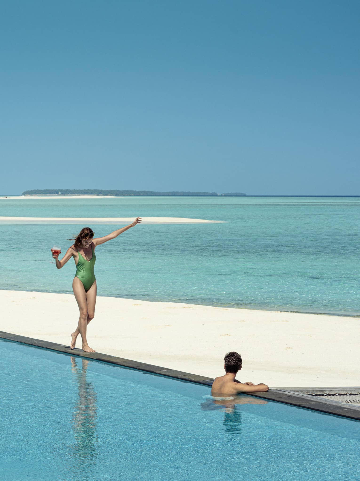 A guest wades in a pool on a beach overlooking the ocean as another guest balances on the edge with a drink in hand.