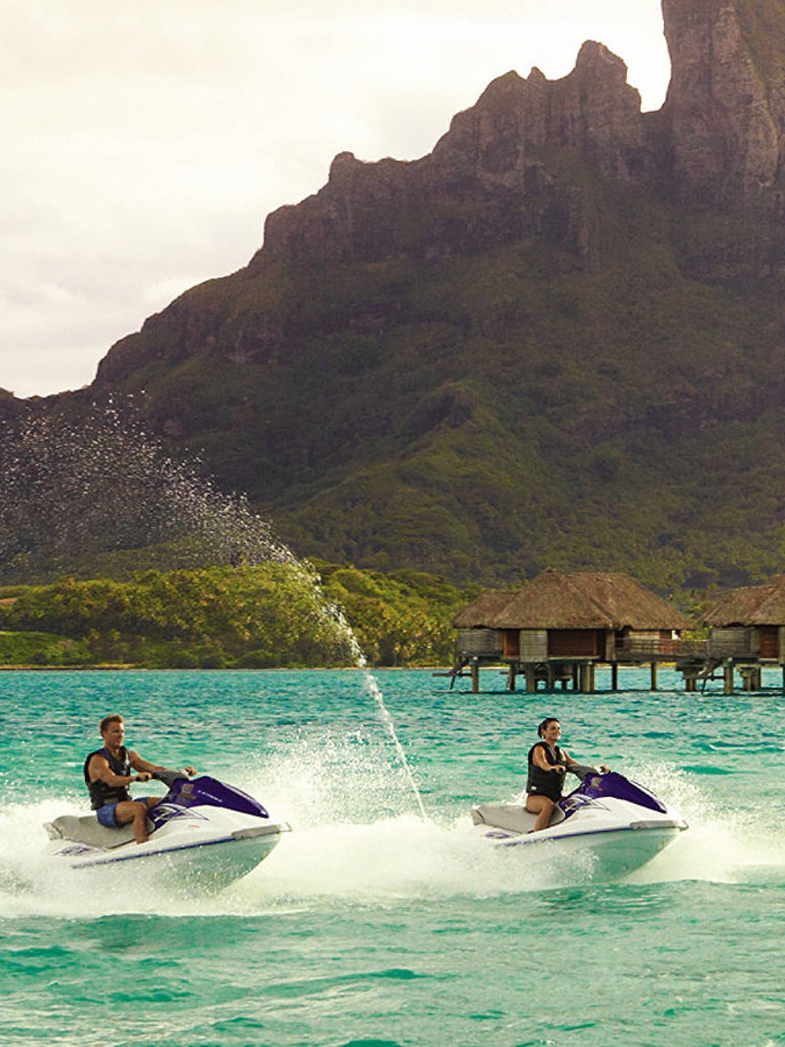 Two jet skis ride along lagoon past row of overwater bungalows, Bora Bora mountain