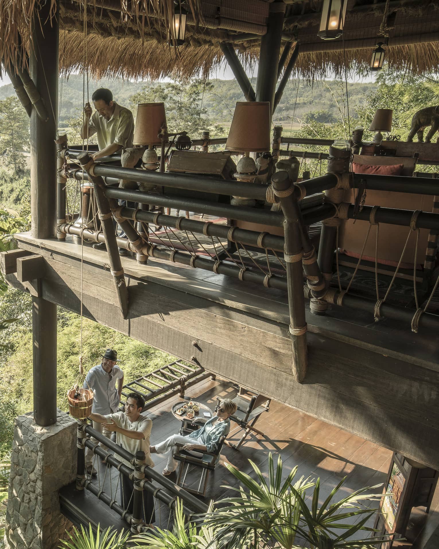 People relaxing on the lower level of a two-storey open-air wooden deck as person passes basket of snacks down on rope