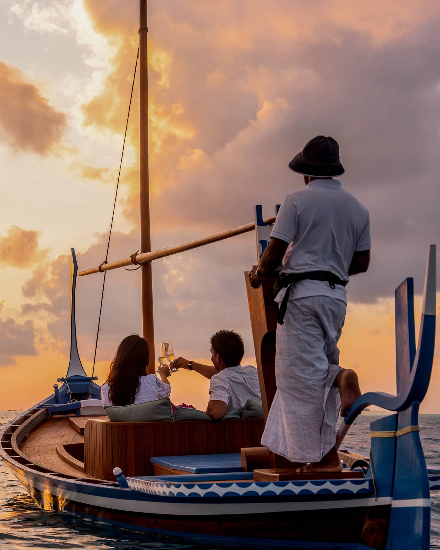 Rear view of two people seated in a dhoni boat, toasting with champagne, the boat handler steering into the golden sunset.