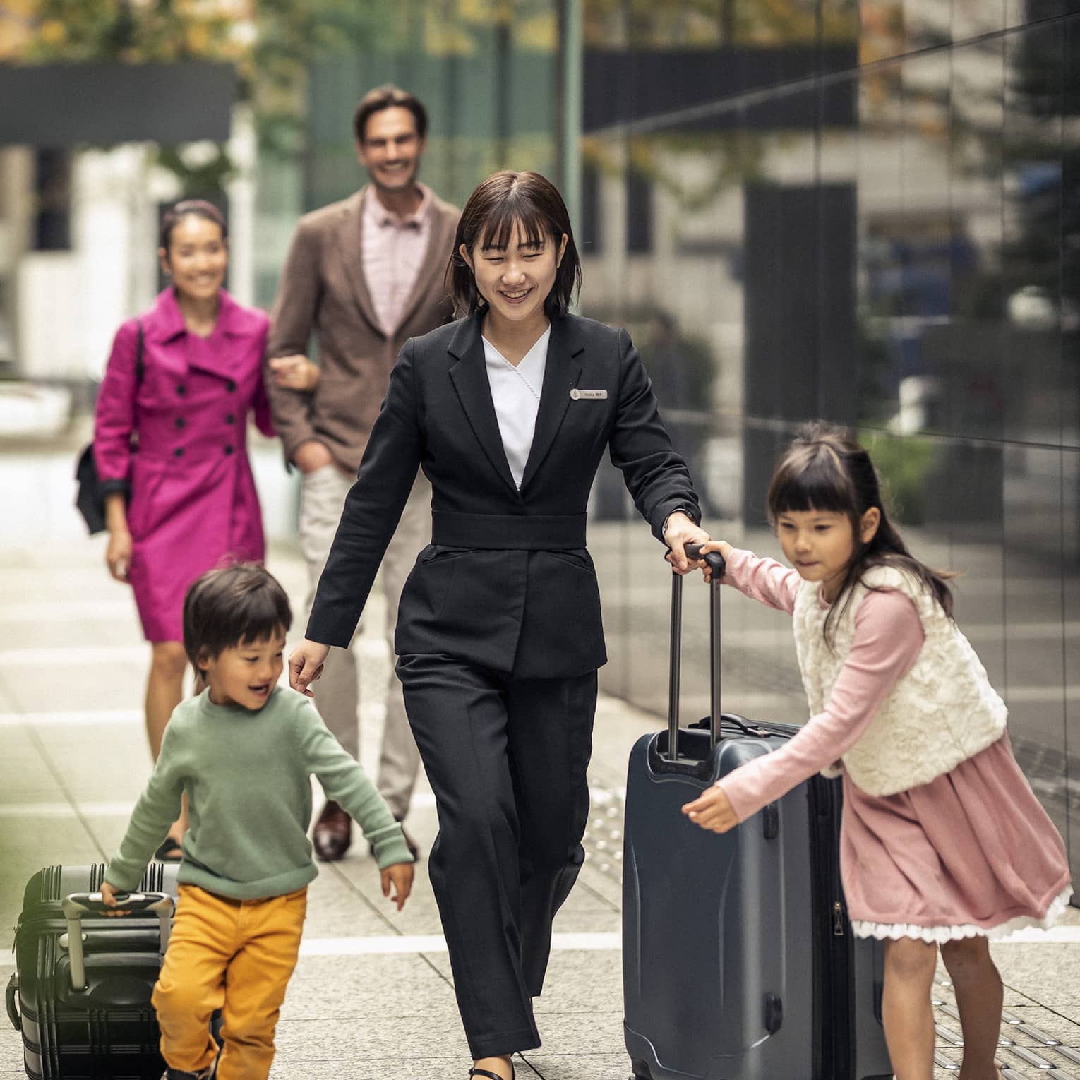 A concierge helps two children wheel their suitcases down a tiled hallway; a couple arm-in-arm follows behind.