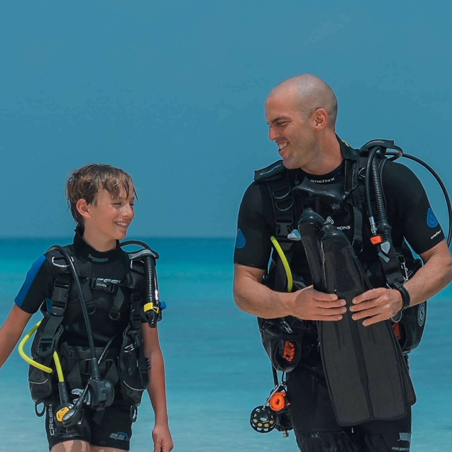 Smiling child and scuba instructor in wetsuits and scuba gear walk at water's edge, aqua ocean behind, cloudless sky above.