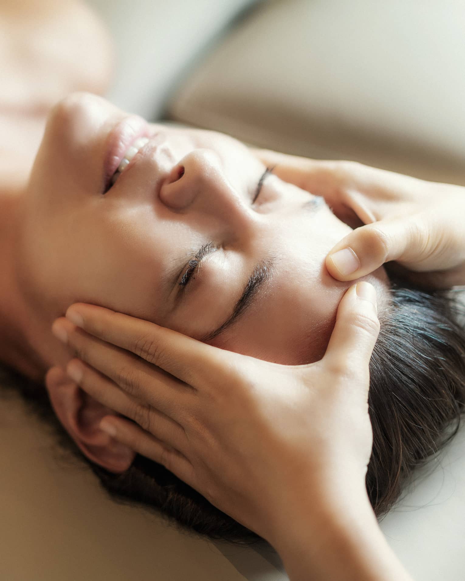 Spa facial, two hands rest on woman's forehead as she closes her eyes, lays under sheet on treatment table