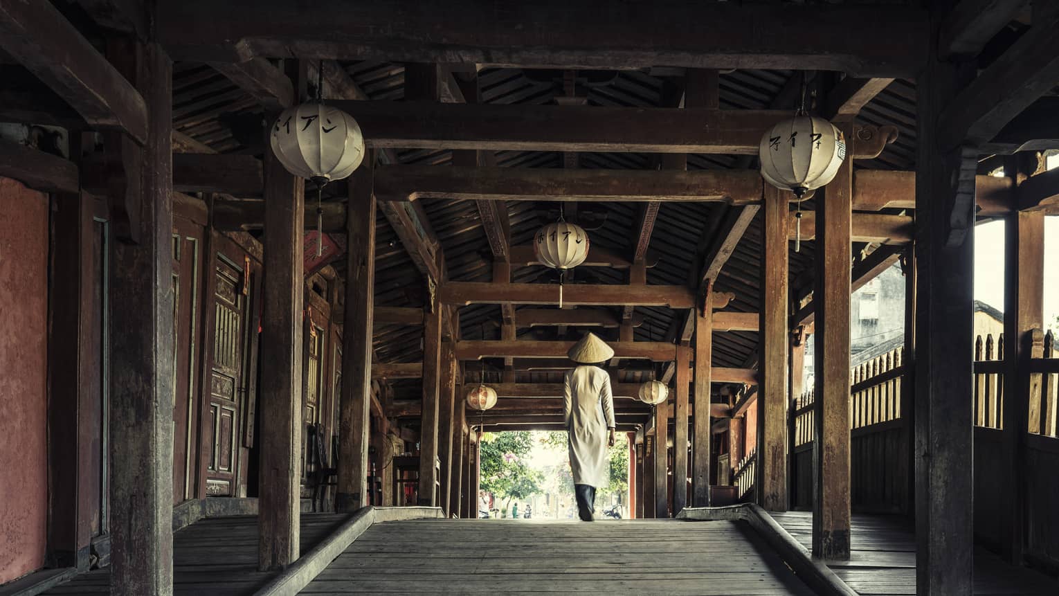 A person wearing traditional Vietnamese attire and a conical hat walks through an ancient wooden outdoor hallway decorated with lanterns.