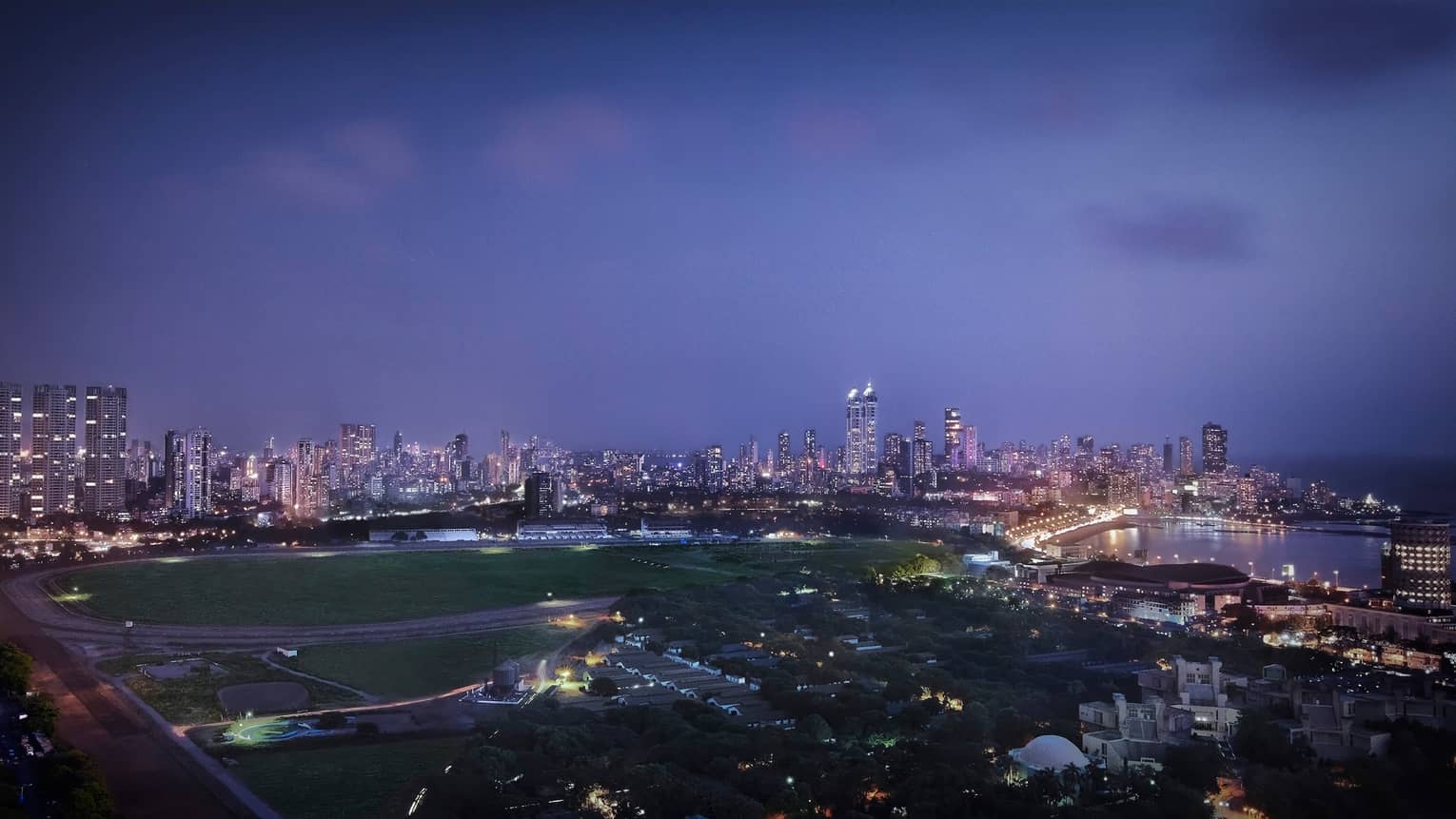 View over large field, track, Mumbai city skyline with buildings, lights at dusk