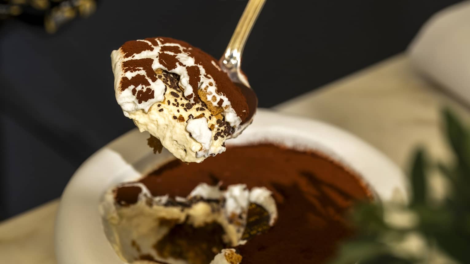 A person spoons a bite of tiramisu out of a around white bowl sitting next to a small cup of coffee.