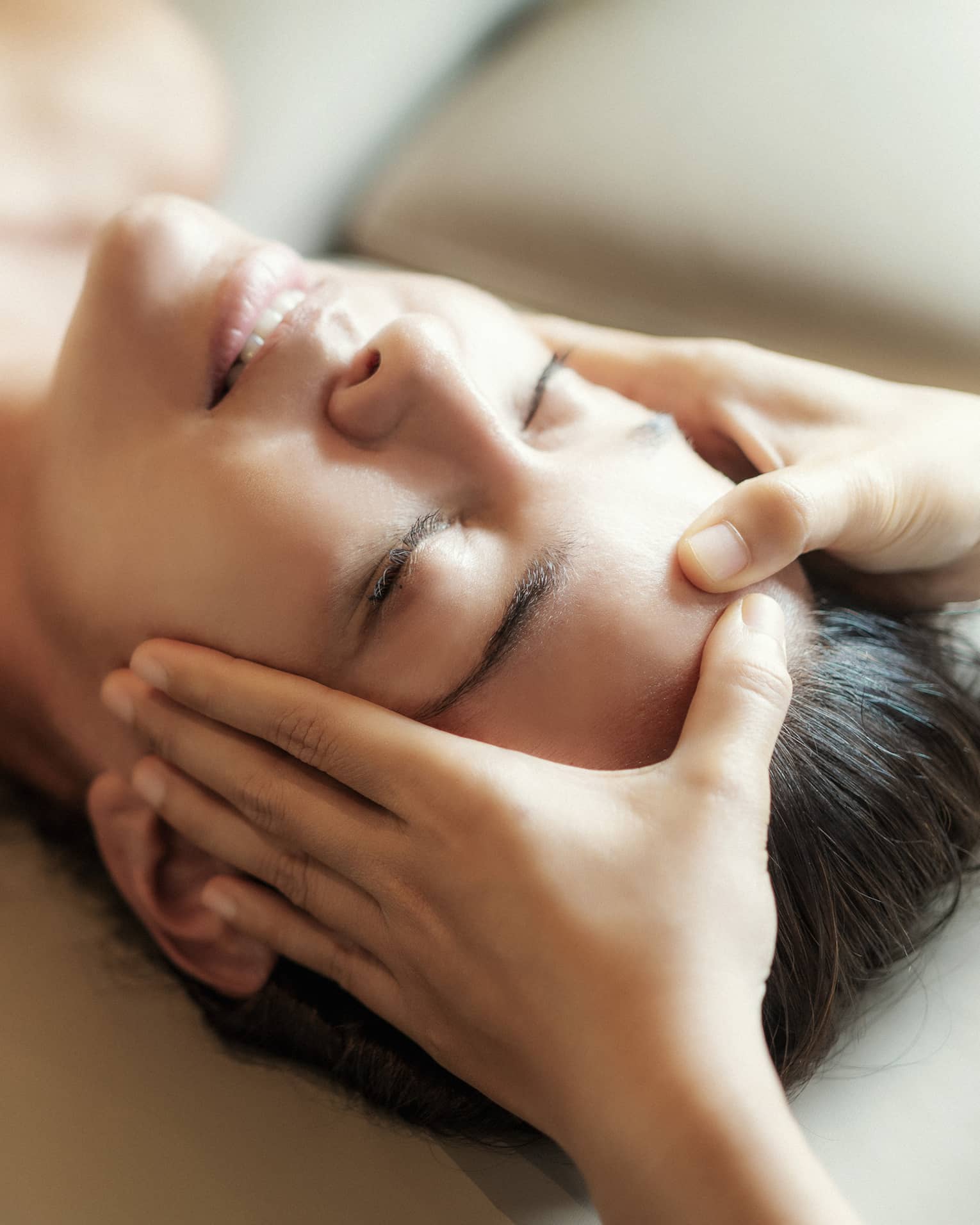 Spa facial, two hands rest on woman's forehead as she closes her eyes, lays under sheet on treatment table