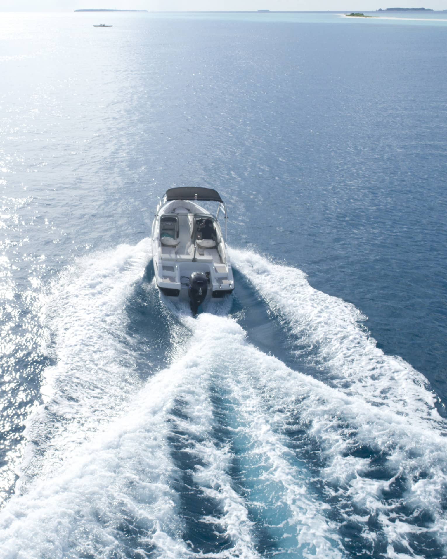 Aerial view of a motorboat towing a wakeboarder, churning up white waves as it speeds through calm water. White sky overhead.