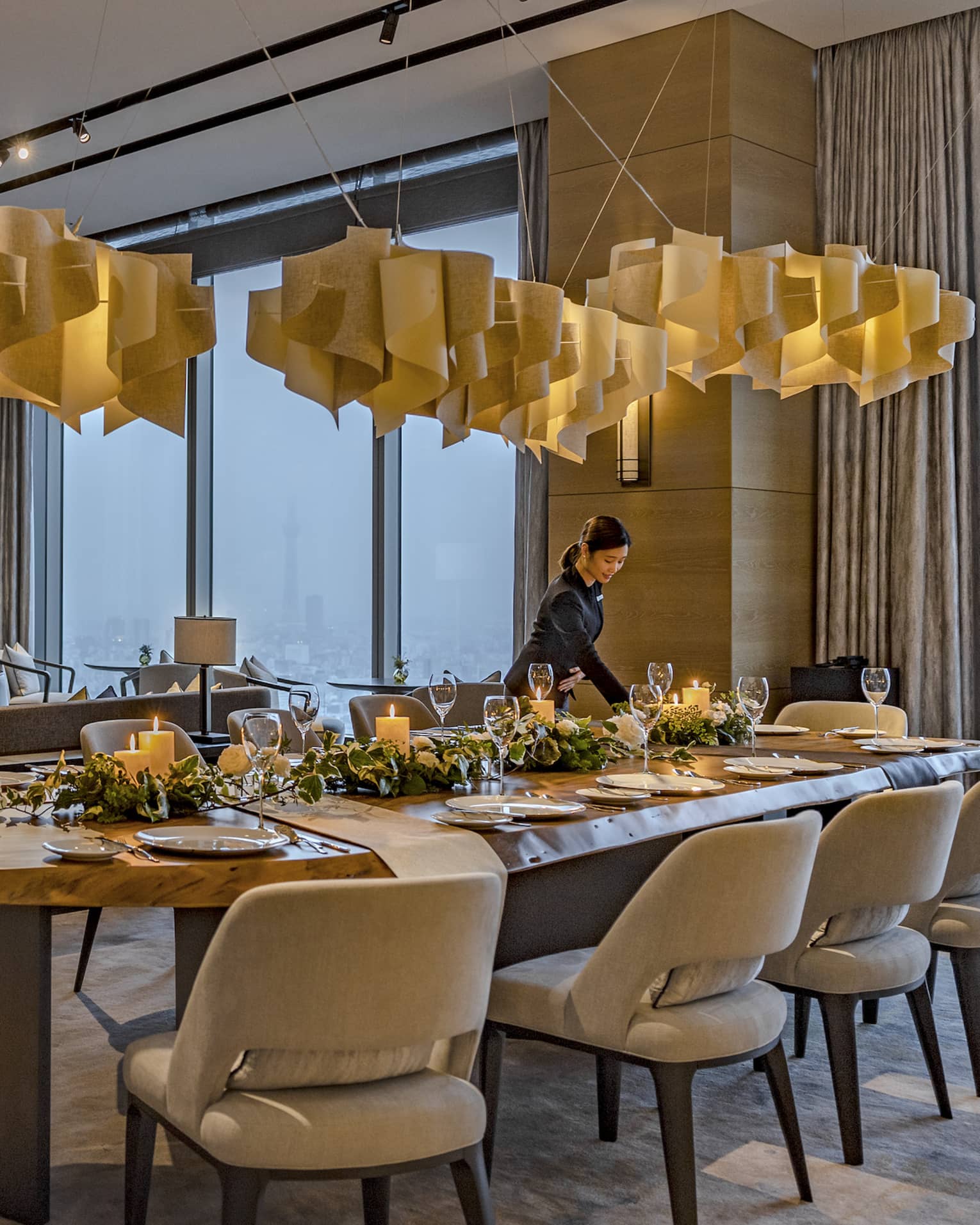 A candlelight dinner is set at a long, oval table with plush seating for twelve under pendant lamps made of swirling wood.