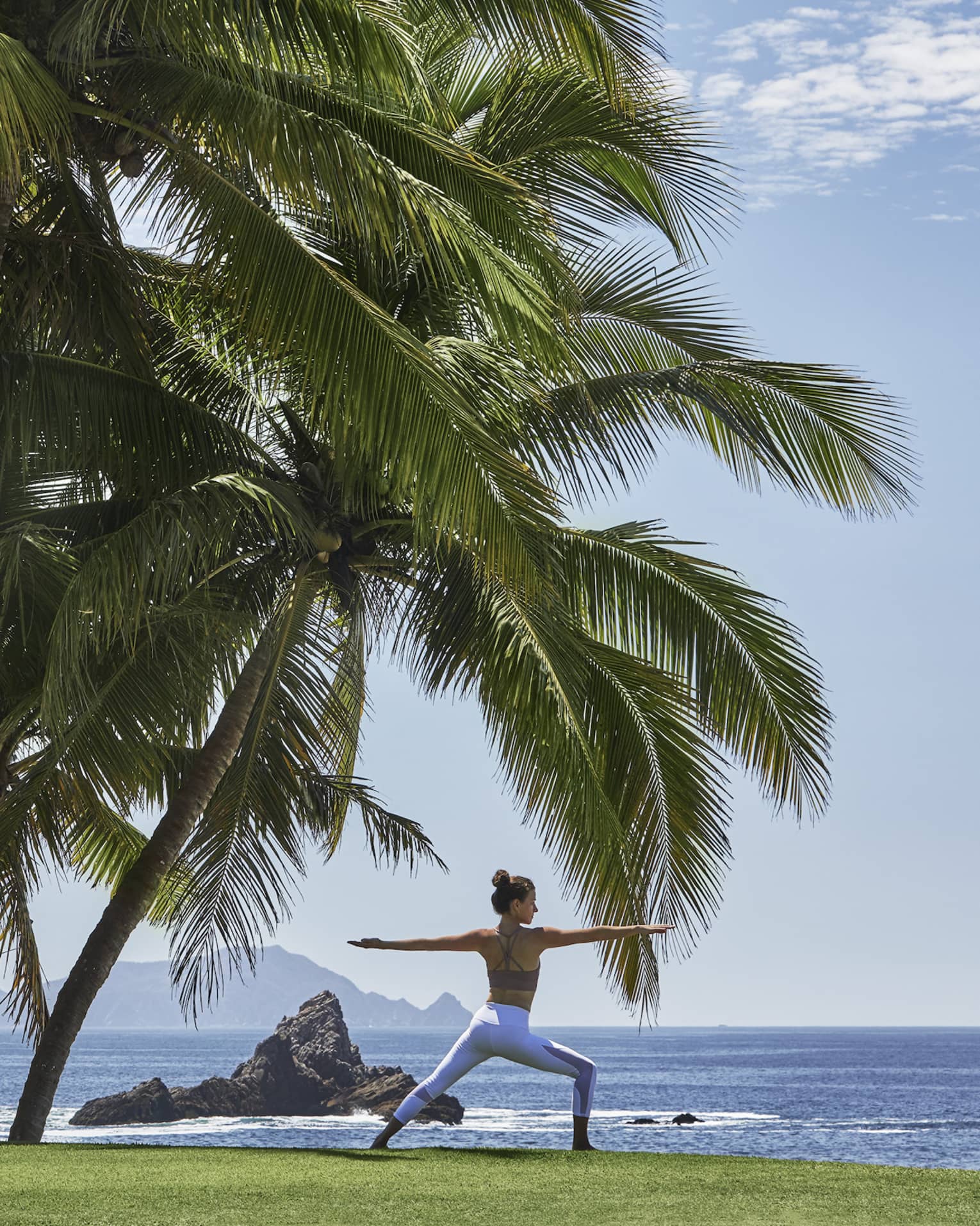 A guest in warrior two yoga pose, dwarfed by a nearby group of immense palm trees, along a sea shore dotted by rock spires. 